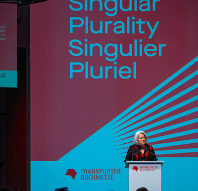 La gouverneure générale Mary May Simon est debout derrière un podium gris. Elle parle dans un microphone. Sur le podium, on peut lire en rouge : «Frankfurter Buchmesse». Derrière elle, un écran affiche «Singular Plurality, Singulier Pluriel».