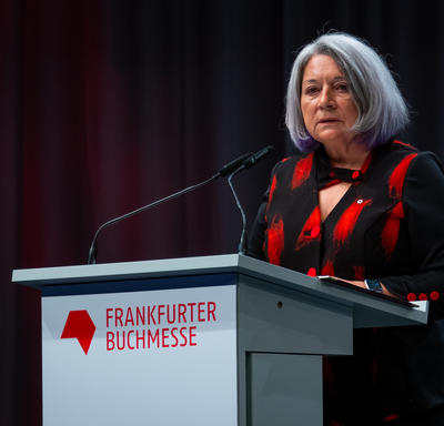 La gouverneure générale Mary May Simon est debout derrière un podium gris. Elle parle dans un microphone. Sur le podium, on peut lire en rouge : «Frankfurter Buchmesse». Elle porte une chemise noire et rouge.