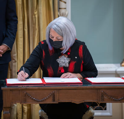 Governor General Mary May Simon is sitting at a desk signing a book.