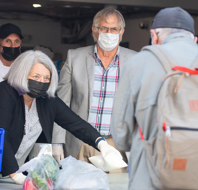  Their Excellencies are serving a meal to a client of the Ottawa Mission. There is a staff member behind them. They are outside and wearing masks.