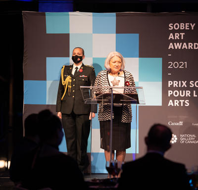 La gouverneure générale est debout sur un podium lors de la cérémonie de remise des Prix Sobey pour les arts 2021. Un membre de l'armée se tient derrière elle.
