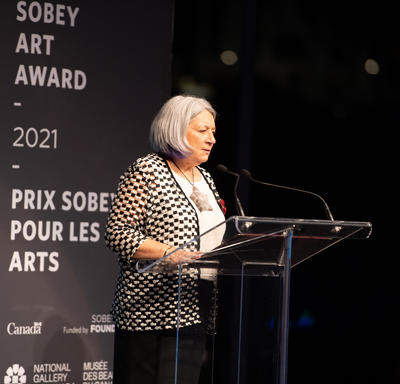 La gouverneure générale est debout sur un podium lors de la cérémonie de remise des Prix Sobey pour les arts 2021. Elle porte un grand collier à pendentif étoile.
