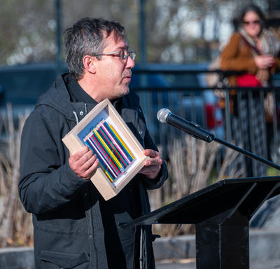 A man speaks at a podium. He is holding a photo box that has a row of crayons in it.