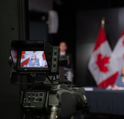 Une caméra enregistre la gouverneure générale Mary Simon. On la voit sur l'écran LCD de la caméra. Elle est également à l'arrière-plan de la photo, hors champ. Derrière elle, on aperçoit des drapeaux canadiens.