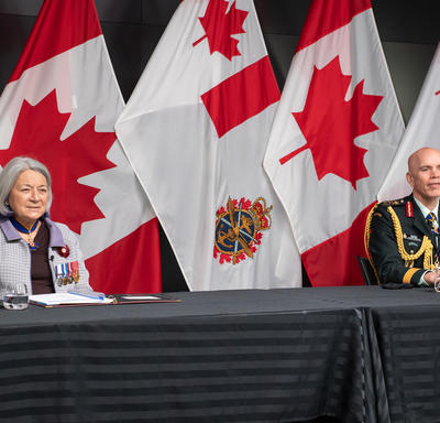 Governor General Mary Simon is sitting at a long table with General Wayne Eyre. They are both facing the camera. Behind them are Canadian and Canadian Armed Forces flags. 