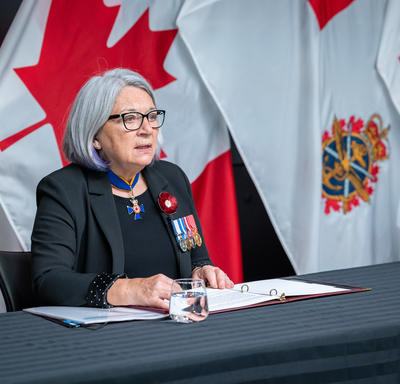 Governor General Mary Simon is sitting at a table. A notebook is open in front of her. The flag of Canada and the flag of the Canadian Armed Forces can be seen behind her.
