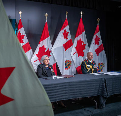 Governor General Mary Simon is sitting at a long table with General Wayne Eyre. They are both facing the camera. Behind them are Canadian and Canadian Armed Forces flags. 