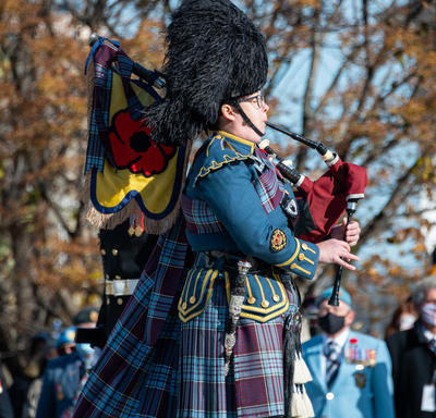A bagpiper plays the bagpipes by the National War Memorial. They are wearing a bearskin hat, and a plaid military uniform. Veterans can be seen in the background.