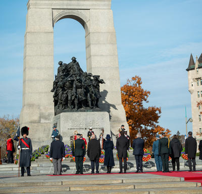 Une file composée de plusieurs personnes fait face au Monument commémoratif de guerre du Canada. La gouverneure générale Mary Simon et M. Whit Fraser se tiennent parmi eux. Le ciel est bleu.