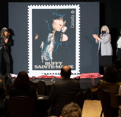 Large stamp unveiling featuring Buffy Sainte-Marie. The Governor General and Buffy Sainte-Marie clap and stand on either side of the display.