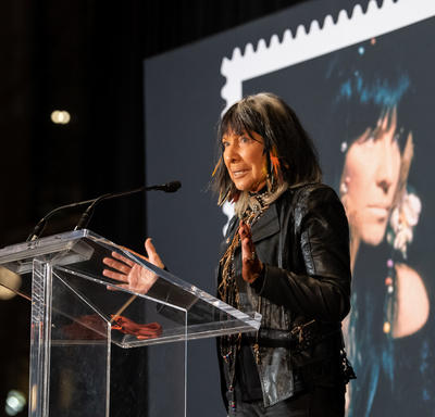 Buffy Sainte-Marie speaks at a podium. A photo of her new commemorative stamp is visible behind her.
