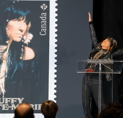 Buffy Sainte-Marie s'exprime sur un podium. Elle célèbre en levant les bras. Une photo de son nouveau timbre commémoratif est visible à sa droite.