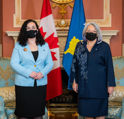La gouverneure générale et la présidente du Kosovo se font photographier. Un drapeau du Canada et un drapeau du Kosovo sont visibles derrière elles. Toutes deux portent un masque.