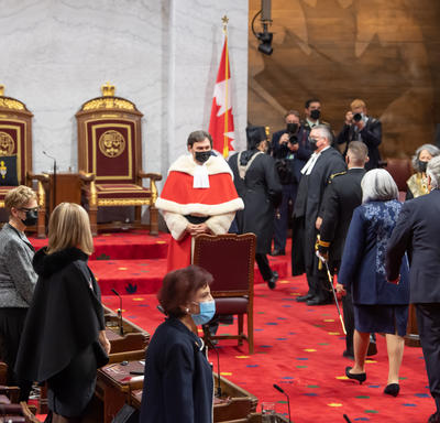 A procession, including the Governor General, walks into the Senate. The Chief Justice of Canada watches as they enter.