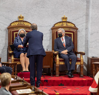 Their Excellencies seated at a pair of thrones. The Secretary to the Governor General presents her with a binder containing the Throne speech.