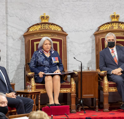 Leurs Excellences sont assises à une paire de trônes. Son Excellence tient le classeur contenant le discours du Trône. Le Premier ministre est à sa droite.