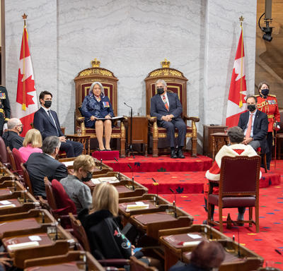 Vue large de la salle du Sénat. Leurs Excellences sont assises dans une paire de sièges trônes, le premier ministre est à la droite de Son Excellence. Le juge en chef du Canada est assis face à leurs Excellences.