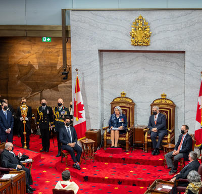Vue large de la salle du Sénat. Leurs Excellences sont assises dans une paire de sièges trônes, le premier ministre est à la droite de Son Excellence. Le juge en chef du Canada est assis face à leurs Excellences.