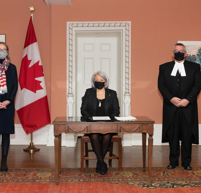 La gouverneure générale se tient entre quatre personnes. Toutes les personnes portent des masques. Un drapeax du Canada se trouve à l’arrière-plan.