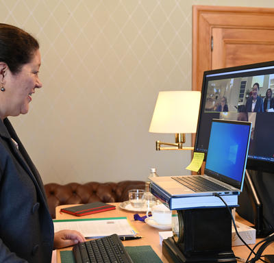 A woman participates in a virtual call on her computer. She is sitting at a desk with two monitors before her.