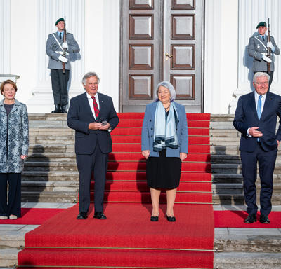 Their Excellencies and two other people are standing on the red carpet at the base of the stairs outside a large white building.