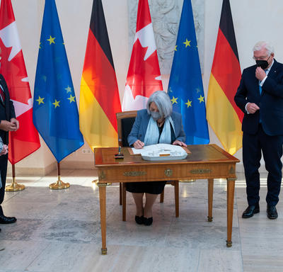 The Governor General is sitting at a wooden desk. She is signing the guest book. President Steinmeier is standing to her left. Ms. Elke Büdenbender and Mr. Whit Grant Fraser are standing to her right. There are several flags behind her. 