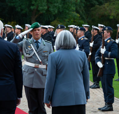 The Governor General and President of Germany are walking on a red carpet at Schloss Bellevue. There are people in military uniform standing along the red carpet.