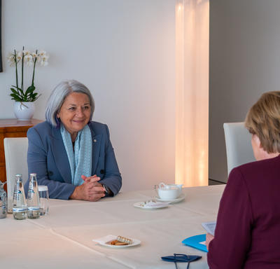 Governor General Mary Simon is sitting at a table. She is sitting across from Her Excellency Angela Merkel, German Chancellor.There is water, warm drinks and pastries on the table. The Governor General is smiling.