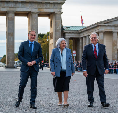 Mr. Michael Müller, the Governing Mayor of Berlin, Governor General Mary Simon and Mr. Whit Grant Fraser. They are smiling and standing on the street in front of the Brandenburg Gate. There is a crowd gathered along the street behind them.