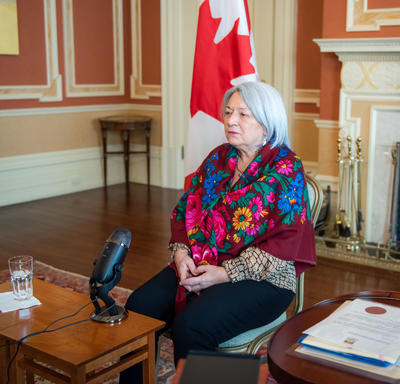 Governor General Mary Simon is sitting down. She is wearing a beautiful multi-coloured scarf. A Canada flag is behind her. A microphone is on a low table in front of her.