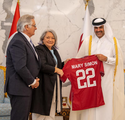 La gouverneure générale Mary Simon et Son Altesse le cheikh Khalid bin Khalifa bin Abdulaziz Al Thani tiennent un chandaille rouge sur lequel est inscrit « Mary Simon 22 ». M. Whit Fraser est debout à côté d’eux.