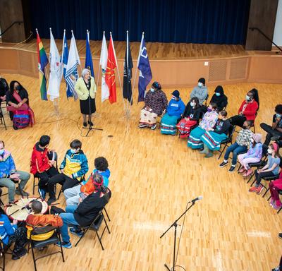 An aerial view of a school gym. Governor General Simon and students are forming a circle. Governor General Simon is standing and speaking to the students.