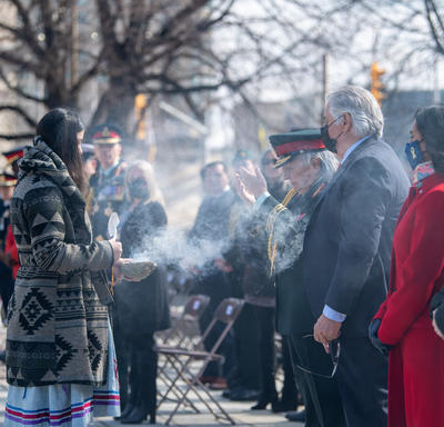 Governor General Simon is standing in a row of people. She is shrouded in smoke from a smudging ceremony. An Indigenous woman performing the smudging ceremony is standing before her.