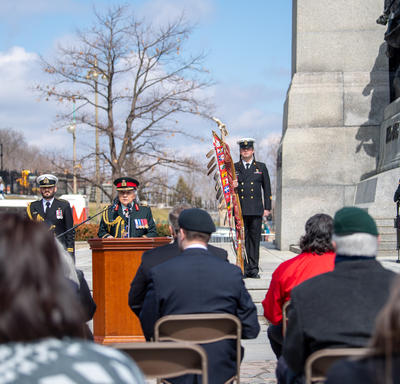 Governor General Simon is delivering remarks at the National War Memorial. People in various military uniforms are standing behind her. The photo is taken from the back of a crowd looking at her.