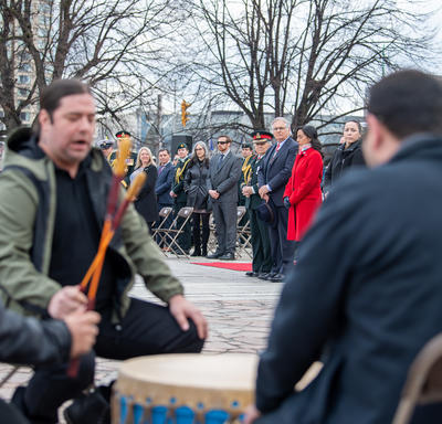 Three men are beating a drum in the forefront of the photo. A crowd in the background of the photo is looking at them. Governor General Simon and Mr. Fraser are standing at the front of the crowd. 