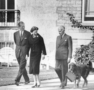 A black-and-white photo of The Queen and The Duke of Edinburgh walking outside Rideau Hall with Governor General Massey. A dog walks alongside them, carrying The Queen’s purse in its mouth. 