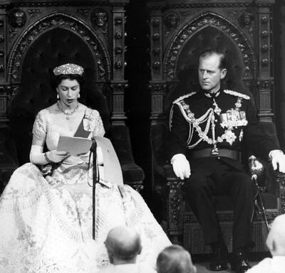 Photo en noir et blanc de Sa Majesté la Reine et du duc d’Édimbourg assis chacun sur un trône dans la salle du Sénat de l’édifice du Parlement. La Reine lit un discours et le duc d’Édimbourg la regarde. Ils sont tous deux en tenue de soirée.