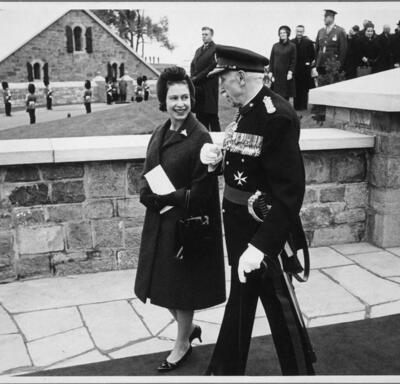 A black-and-white photo of The Queen walking with Governor General Vanier. They are walking along the stone ramparts of the Citadelle. A small crowd of people, guards in uniform and another stone building are in the background.