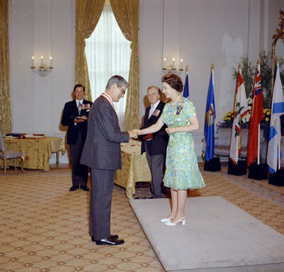 Jules Léger, dressed in a grey suit, shakes the hand of The Queen in the Ballroom at Rideau Hall. The Queen stands in front of him in a green dress and white heels. Three men look on in the background.