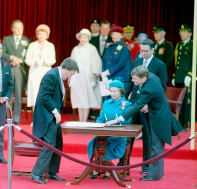 La Reine, vêtue d’un manteau et d’un chapeau bleus, est assise à une table en bois. Un grand document est posé sur la table. Plusieurs politiciens et autres personnes sont debout, sur un grand tapis rouge, et regardent la scène.