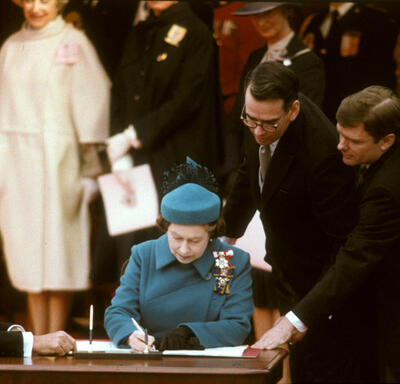 The Queen, dressed in a blue coat and hat, is seated at a wooden table. She is signing a document. Then-Prime Minister Pierre Elliott Trudeau is also seated at the table. Three other men look on.  