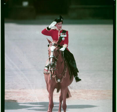 La Reine, vêtue d’un uniforme, monte son cheval de côté. Elle porte des gants blancs et salue de la main droite.