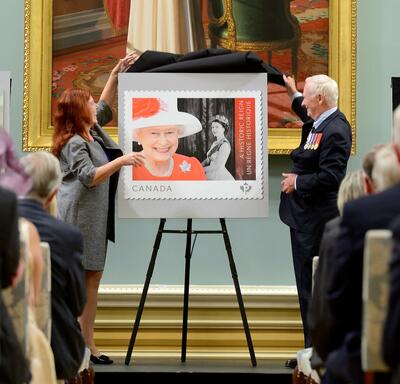 Le gouverneur général de l’époque, David Johnston, et Mme Siân Matthews, de Postes Canada, dévoilent un nouveau timbre en l’honneur de la Reine. Le timbre présente une image en couleur de Sa Majesté et une ancienne photo en noir et blanc de sa jeunesse.