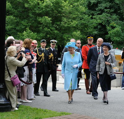 The Queen, in a matching blue coat and hat, walks along a pathway surrounded by trees on the Rideau Hall grounds. She is accompanied by the Secretary to the Governor General and by The Duke of Edinburgh. She smiles at a crowd of people. 