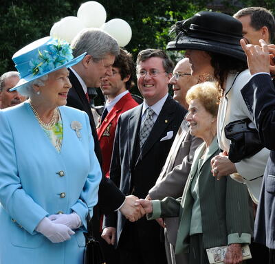 The Queen, in a matching blue coat and hat, smiles at people in a crowd. Behind her, then-Prime Minister Stephen Harper shakes hands with an elderly woman. 