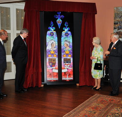The Queen stands beside a stained glass window installment. A set of red curtains hangs on either side of the window. The Queen is accompanied by five other people. 