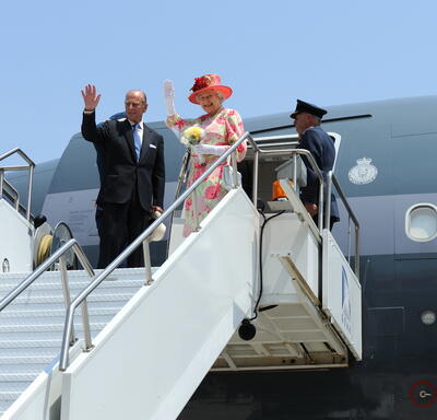 The Queen and The Duke of Edinburgh wave from atop a set of stairs connected to a plane. 