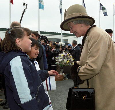 The Queen, dressed in a beige coat and matching hat, receives a bouquet of flowers from two children. A crowd of people stand behind them. Several flags on flagpoles are in the background.