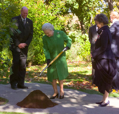The Queen, wearing a green coat, holds a shovel above a small pile of dirt. Then-Governor General Adrienne Clarkson stands to her left. Greenery and trees are in the background.