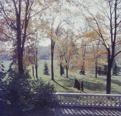 The Queen and then-Governor General Vincent Massey walk together across a lawn surrounded by trees in fall foliage.